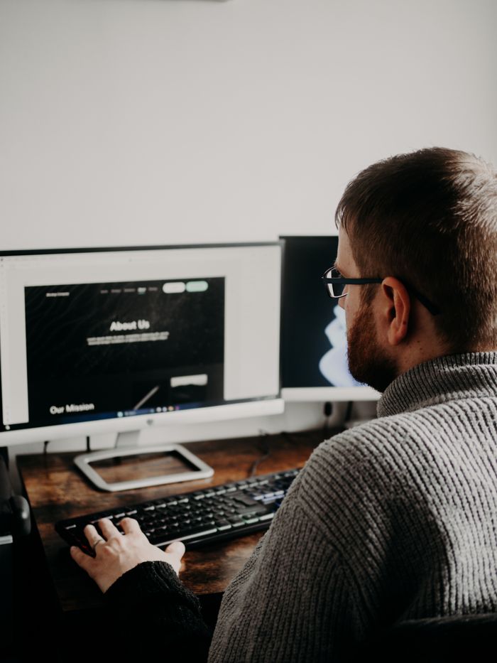 about-us-01 Man working on web design at home office setup with dual monitors.